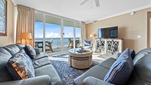 A modern living room with a sofa, a coffee table, and a television. There is a balcony with a view of the ocean.
