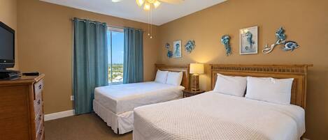A hotel room with two beds, a ceiling fan, a television, and artwork on the wall.