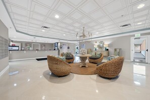 Um lobby com balcão de recepção, cadeiras de vime, uma mesa central e um lustre.