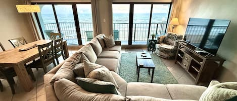 Una sala de estar con un sofá grande, una televisión y un balcón con vistas al mar.
