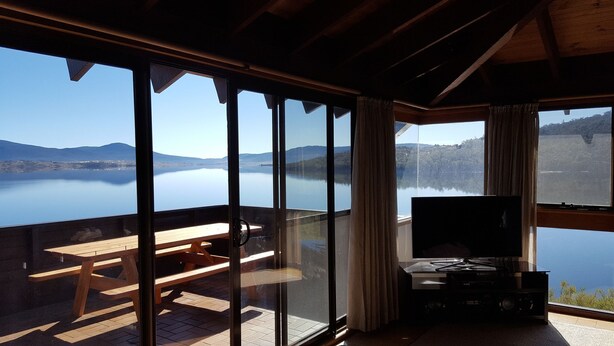 Habitación con grandes ventanales que ofrecen vistas a un lago y montañas. Hay una mesa de madera y un televisor de pantalla plana sobre un soporte.