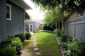 A residential backyard with a stone pathway leading to a house entrance, surrounded by lush greenery and a wooden fence.