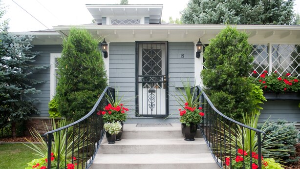 A house with a gray exterior, a black door with a decorative pattern, and a number '15' on the wall. There are two potted plants with red flowers on the steps. - Beautiful Historic Old Northend Bungalow with amazing outdoor space (Colorado Springs)