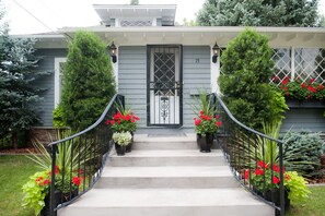 Una casa de fachada gris, una puerta negra con diseño decorativo y el número 15 en la pared. Hay dos macetas con flores rojas en los escalones.