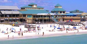 Ein Strandhotel mit mehreren Gebäuden, einer Promenade und einem Sandstrand voller Menschen.