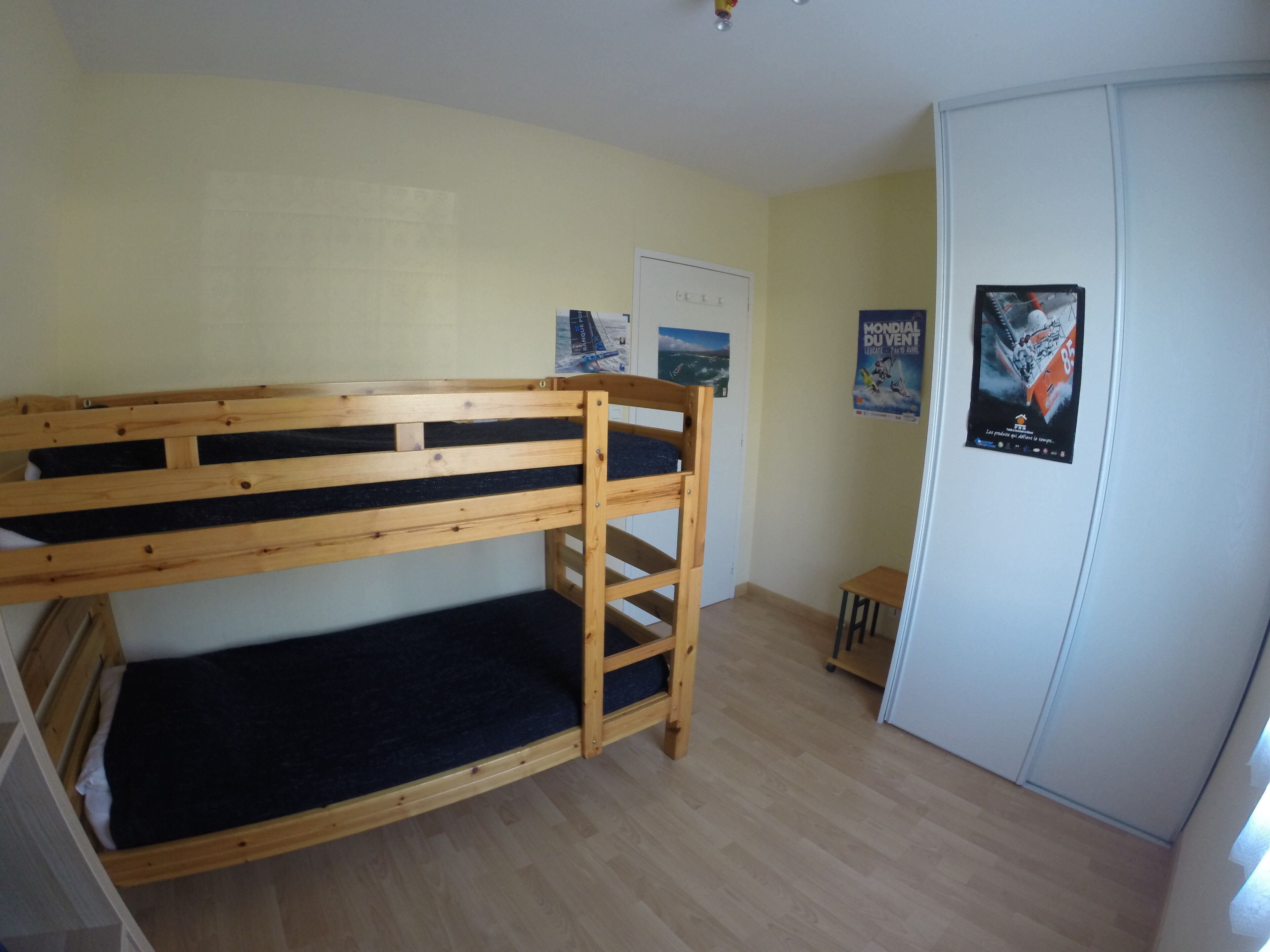 Un lit superposé dans une pièce dont le sol est en bois, avec une armoire blanche et deux affiches accrochées au mur.