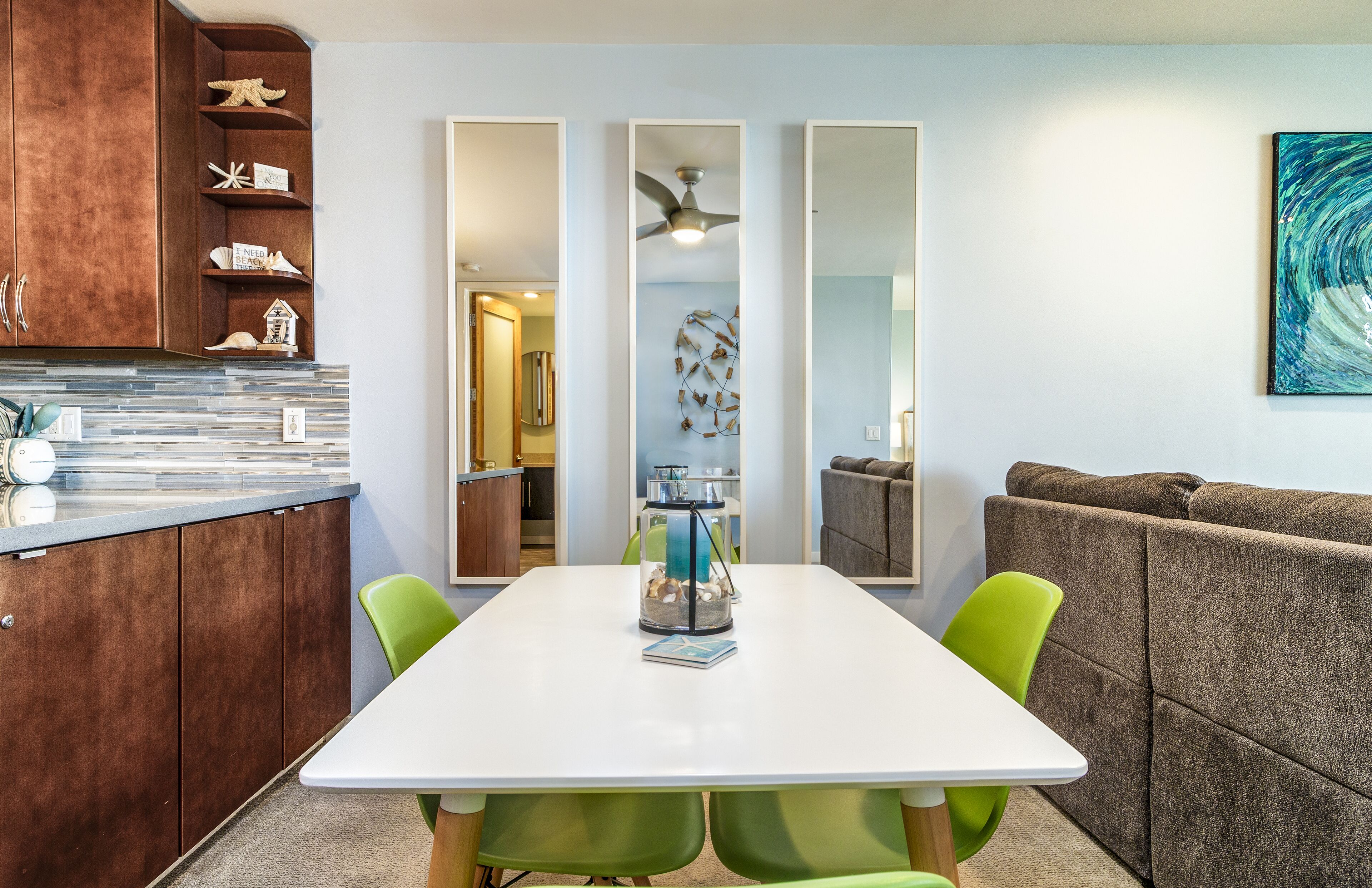 A modern kitchen with wooden cabinets, a white countertop, and a dining table with green chairs. There is a kitchen island with a vase and a book on it.