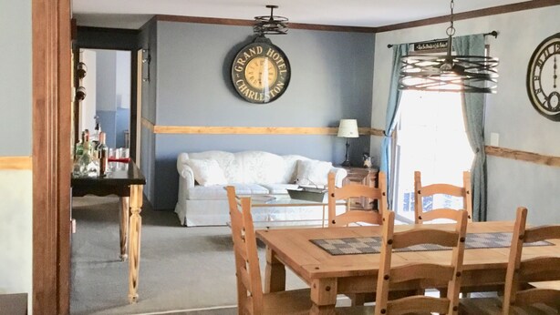 Een eetgedeelte met een houten tafel en stoelen, een klok aan de muur, en een bank op de achtergrond.