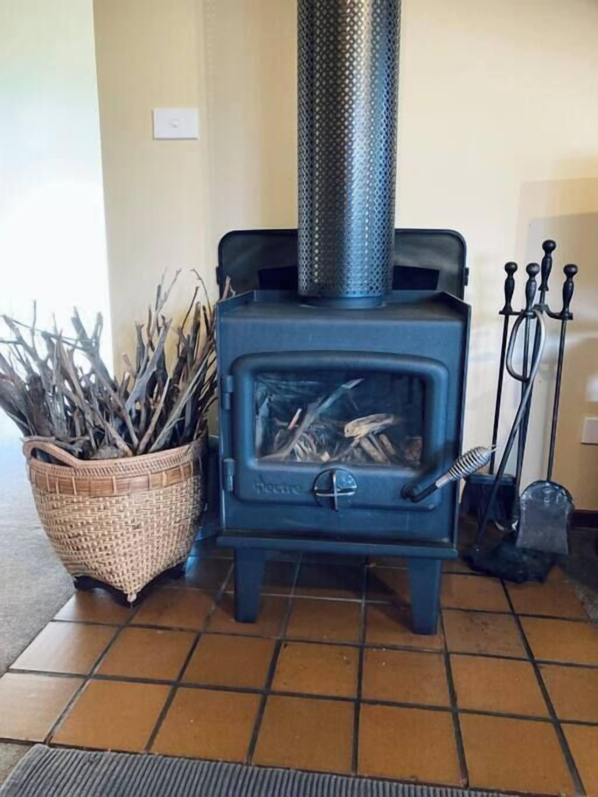 Un poêle à bois bleu avec une cheminée en métal, un panier en osier rempli de bois de chauffage et une brosse en métal.