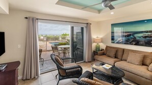 Ein modernes Wohnzimmer mit einer Couch, Sesseln, einem Couchtisch und einem Fernseher. Ein Balkon bietet Blick ins Freie.