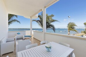 Ein Balkon mit weißen Korbmöbeln, einem Tisch mit einer Vase und Blick auf das Meer mit Booten.