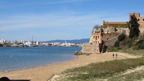 A coastal cityscape with a prominent historical building overlooking the sea, a sandy beach, and a marina with boats.