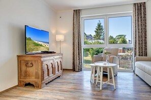 A living room with a television on a wooden cabinet, a round white table with chairs, a sofa, and a large window with a view of a beach and houses.