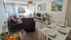 Een moderne woonkamer met een bank, een eettafel gedekt voor twee personen en een televisie. Er staan potplanten en er hangt een schilderij aan de muur.