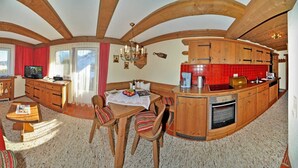 Een gezellige kamer met een bank, een kleine tafel, een televisie en een keukenhoek met houten kastjes en een rode tegelachterwand.