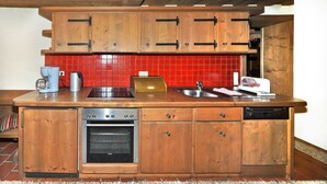 Een compacte keuken met houten kastjes, een inbouwoven, een koffiezetapparaat en een rode betegelde achterwand.