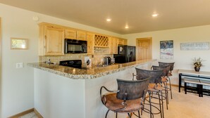 Una cocina con encimeras de granito, armarios de madera, un refrigerador negro y un microondas. Hay taburetes de bar y una mesa de comedor con sillas.