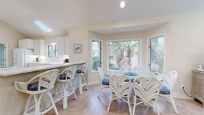 A kitchen with white cabinets, a white refrigerator, a dining area with white chairs and a glass table, and large windows with a view of palm trees. - Comfortable Palmetto Dunes Home with Private Pool and Screened in Porch (Hilton Head)