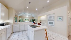 A modern kitchen with white cabinetry, a central island, and a dining area with a view of the outdoors. - Comfortable Palmetto Dunes Home with Private Pool and Screened in Porch (Hilton Head)