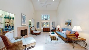 A spacious living room with a fireplace, wicker furniture, and a view through large windows. - Comfortable Palmetto Dunes Home with Private Pool and Screened in Porch (Hilton Head)