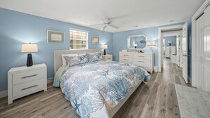 A bedroom with a bed, nightstands, a ceiling fan, and a hallway leading to a bathroom. - Island House Beach Resort V33 (Siesta Key)