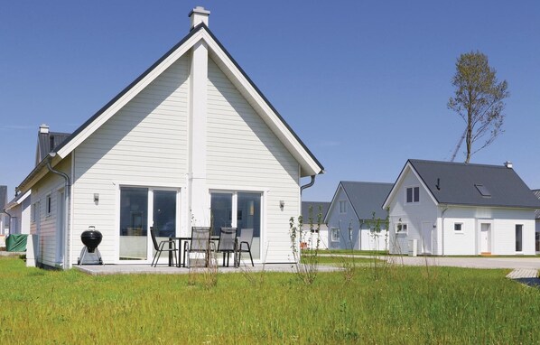 Une maison blanche à deux étages, avec un toit à pignon, un porche à l’avant doté d’un barbecue et un ciel dégagé.