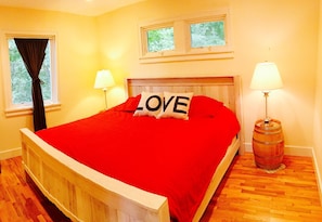 Une chambre à coucher avec un lit en bois, une literie rouge, deux lampes de chevet, une lampe en forme de tonneau en bois et une fenêtre donnant sur des arbres.