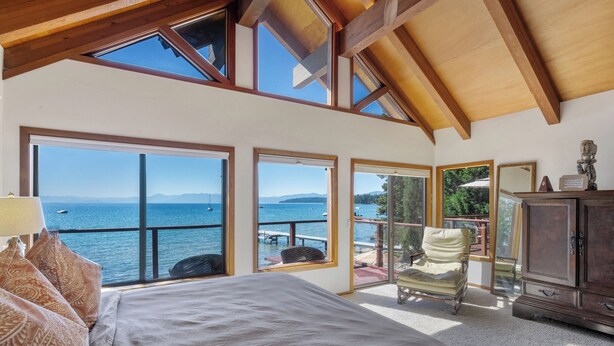 A bedroom with a large bed, a chair, a cabinet, and a view of the sea through large windows. - Lake Front Retreat With Your Private Beach & Hot Tub  (Tahoe Vista)