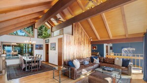 A spacious living area with a high ceiling, wooden beams, a large sofa set, a dining table, and a view of the outdoors. - Lake Front Retreat With Your Private Beach & Hot Tub  (Tahoe Vista)