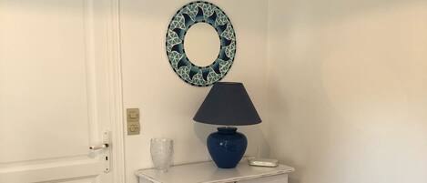Une commode blanche avec des tiroirs, une lampe bleue, une assiette décorative et une chaise blanche dans une pièce dont le sol est recouvert de carreaux marron et qui possède une porte blanche.