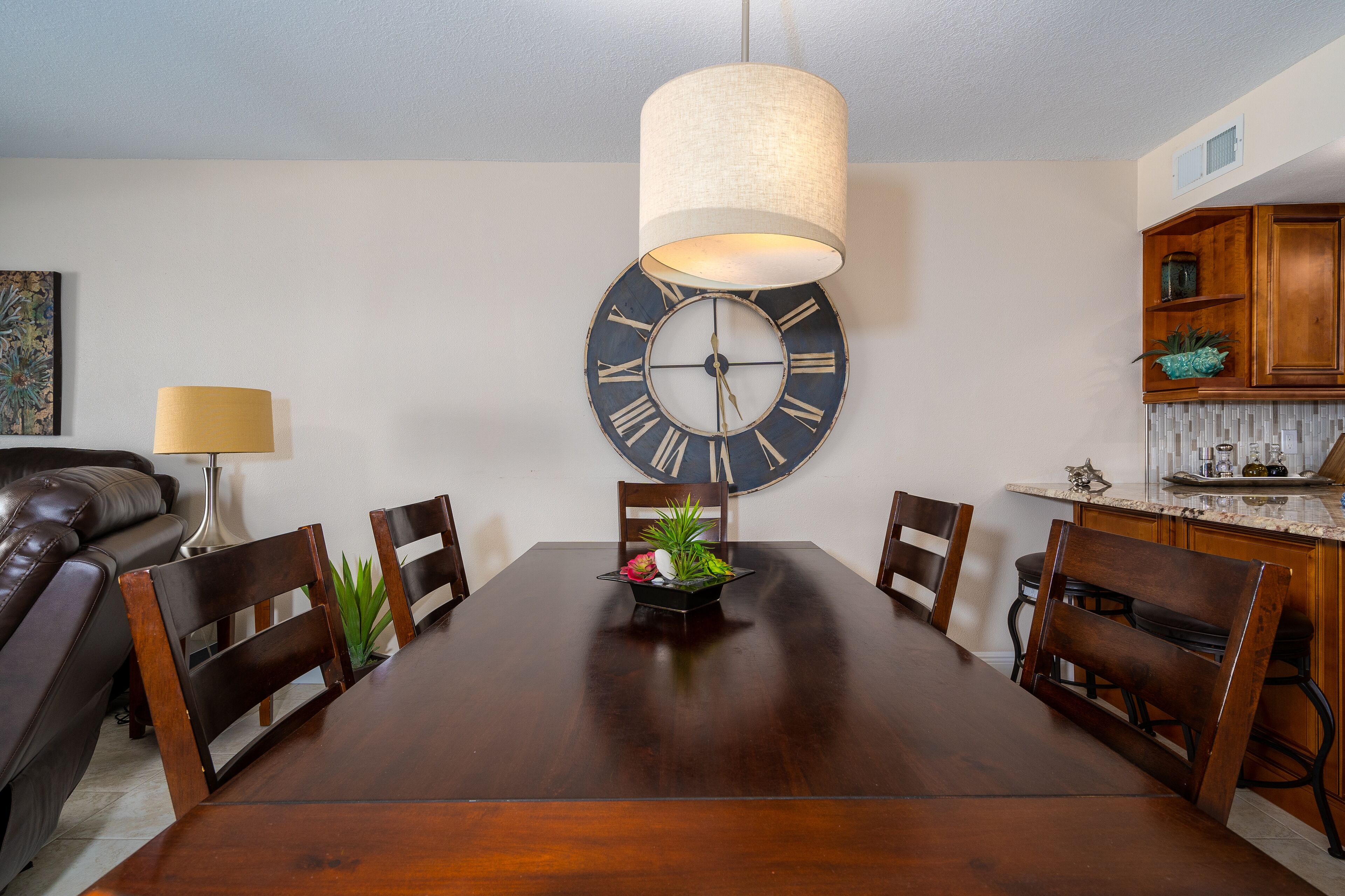 A dining room with a wooden table, chairs, a large wall clock, and a hanging light fixture.