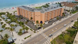Un hotel de varios pisos con vista a la playa, situado cerca de una carretera costera con palmeras y gente disfrutando la playa.