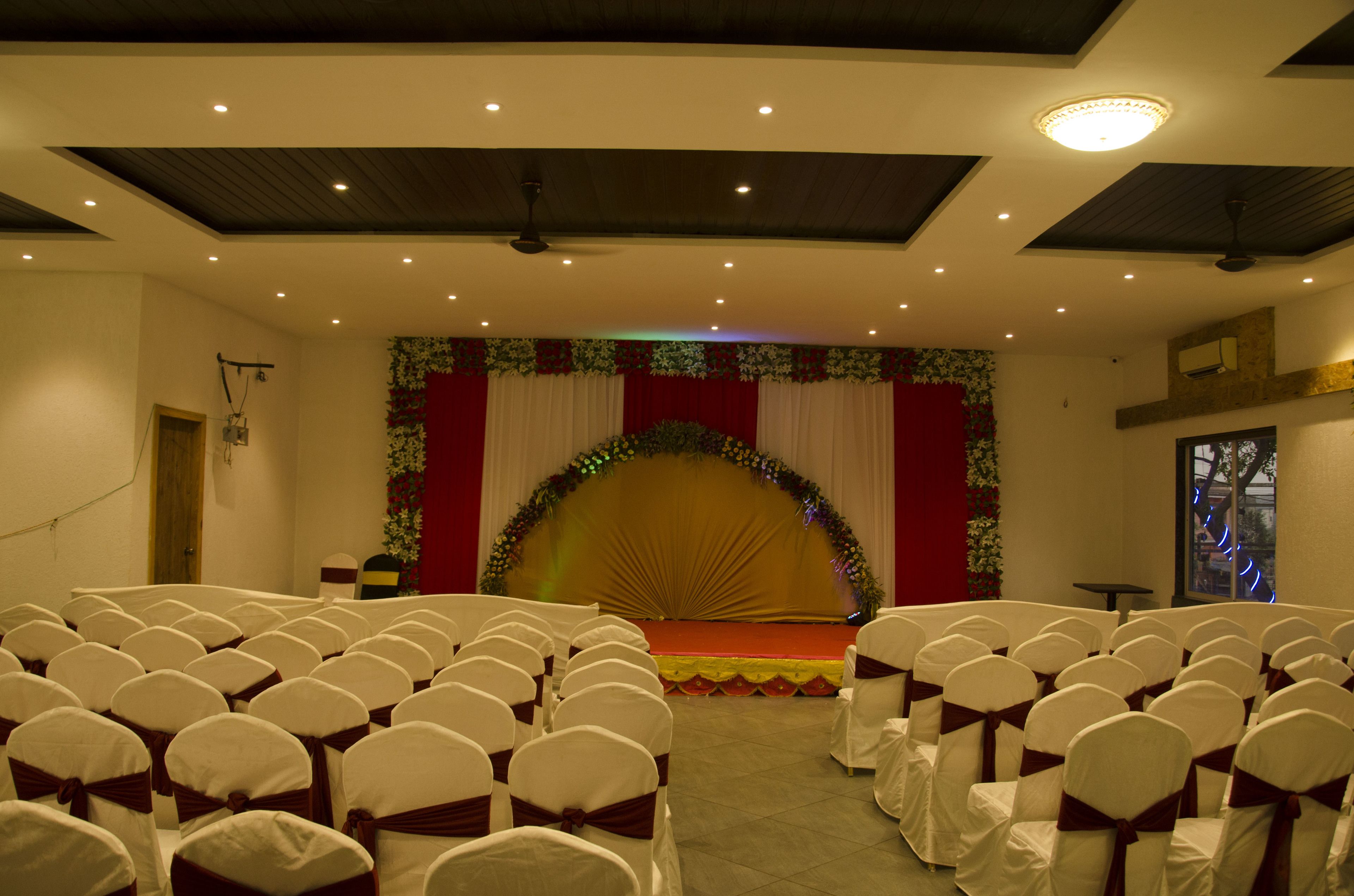 A conference room with rows of white chairs with brown sashes, a stage with a red curtain and floral decorations, and a ceiling with recessed lighting and a fan.