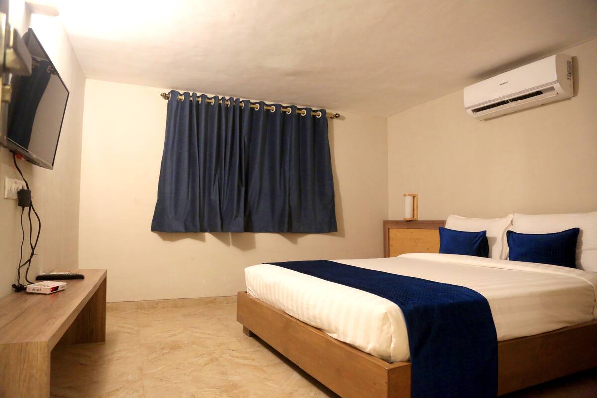 A bedroom with a bed, a TV mounted on the wall, an air conditioner, and a bedside table with a remote control.