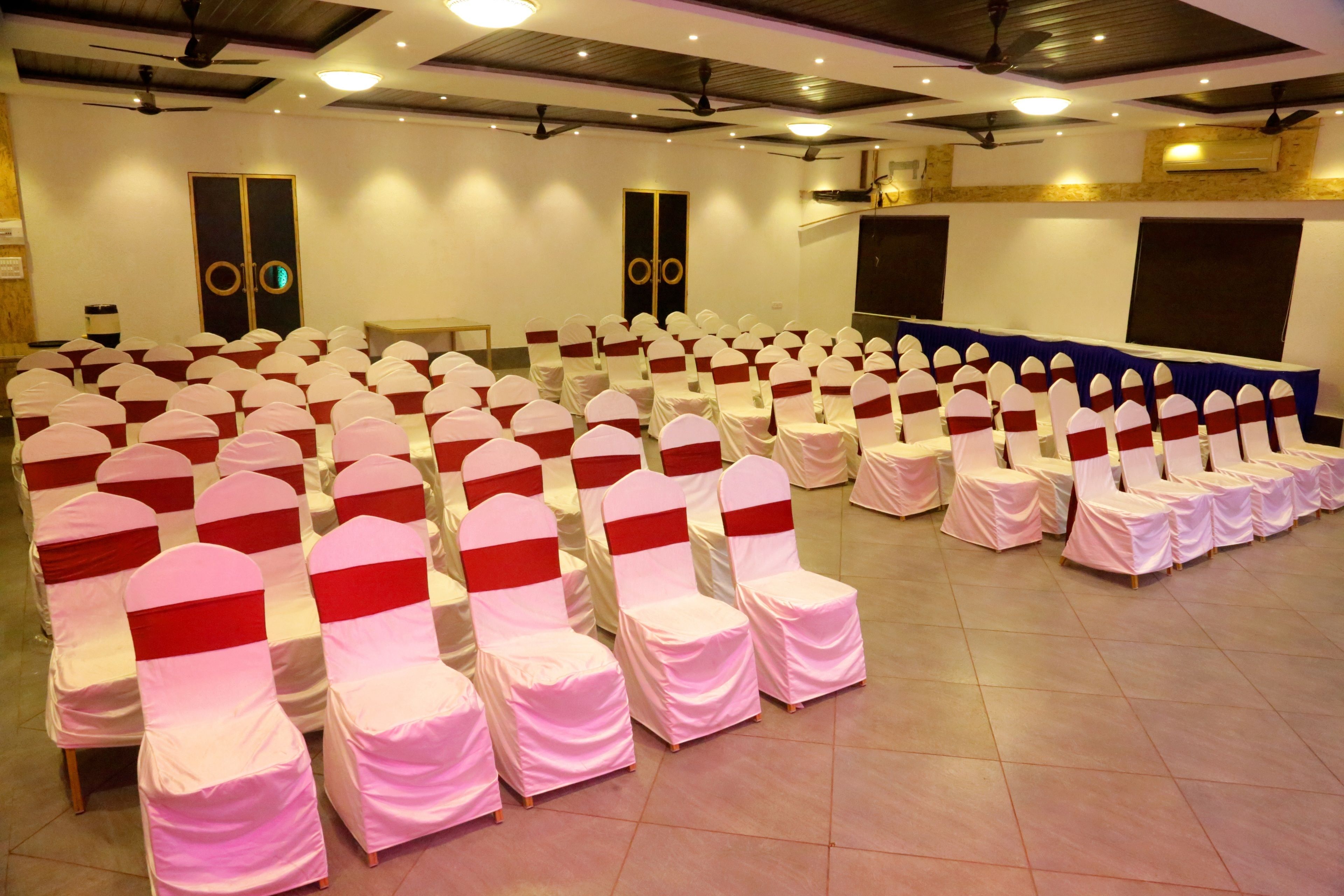 A conference room with rows of chairs covered in white and red sashes, a stage with a blue backdrop, and a ceiling fan.