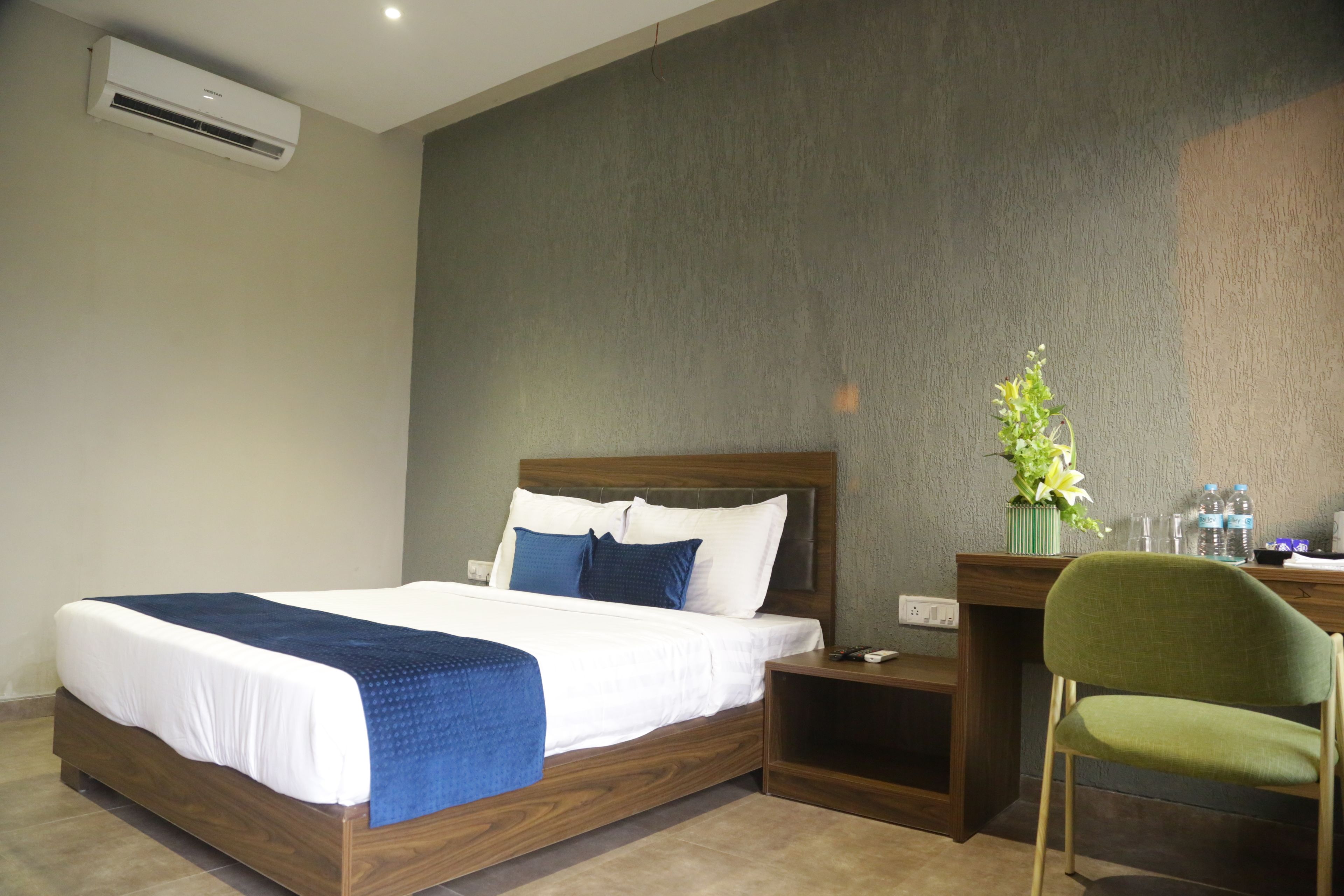 Habitación de hotel con una cama de madera, una silla verde, un escritorio con un florero y un equipo de aire acondicionado en la pared.