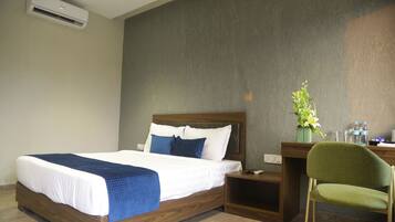 Habitación de hotel con una cama de madera, una silla verde, un escritorio con un florero y un equipo de aire acondicionado en la pared.