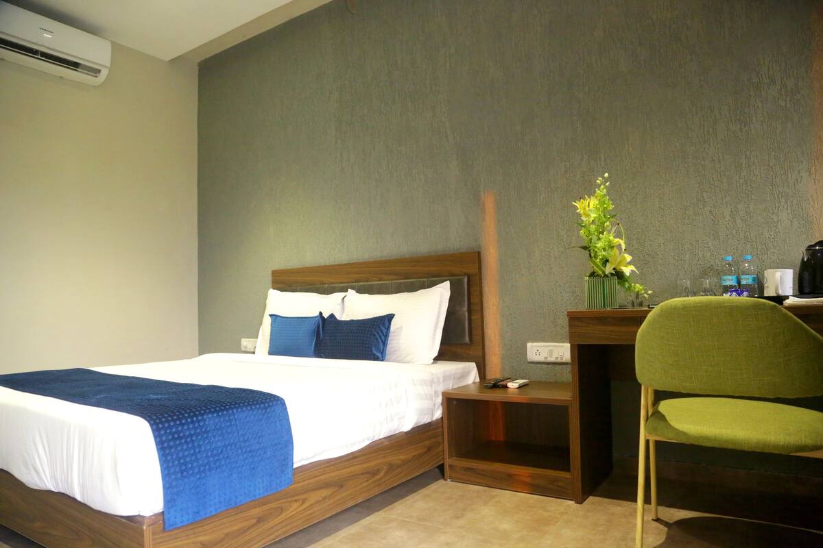 A hotel room with a bed, a desk, a chair, a vase with flowers, and a coffee machine.