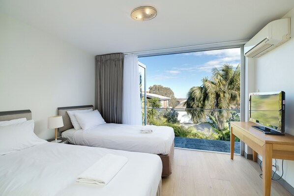 A hotel room with two beds, a TV, and a balcony with a view of palm trees and buildings.
