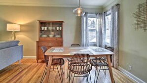 Een eethoek met een houten tafel, vier rieten stoelen, een kast met servies, een lamp en een raam met gordijnen.
