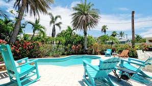 A pool area with turquoise chairs, a small pool, and lush greenery including palm trees and flowering plants. - Poolside Relaxation Just Steps from the Gulf (Anna Maria)