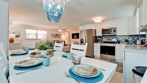 A modern kitchen with white cabinets, a stainless steel refrigerator, a microwave, and a stove. There is a dining area with a white table set for two, blue placemats, and a potted plant. - Poolside Relaxation Just Steps from the Gulf (Anna Maria)