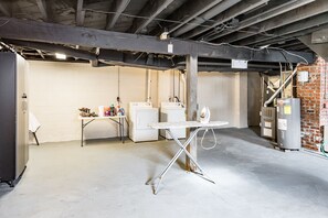 Een kelder met een betonvloer, een tafel op metalen poten, een wasmachine en een boiler.