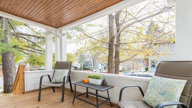 A covered porch with two chairs and a table, a wooden deck, and a view of a street with trees and houses.