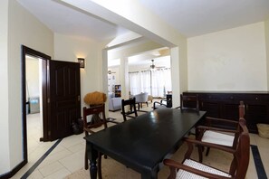 Ein Essbereich mit einem langen Tisch und Stühlen, einer dunklen Holztür und Blick in ein Wohnzimmer mit einer Couch und Vorhängen.