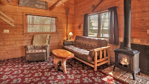 A cozy cabin interior with a wooden floor, a sofa, a chair, a small wooden table, a window with blinds, and a wood-burning stove. - Cabin on AuTrain River a few blocks from Lk Superior, 10 mi from Munising (Au Train)