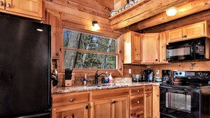 A wooden cabin kitchen with a black refrigerator, wooden cabinets, a granite countertop, a sink, a microwave, a stove, and a window with a view of trees. - Cabin on AuTrain River a few blocks from Lk Superior, 10 mi from Munising (Au Train)