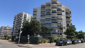 Un immeuble résidentiel à plusieurs étages, doté de balcons, d’un espace de stationnement et surplombant un ciel bleu dégagé.