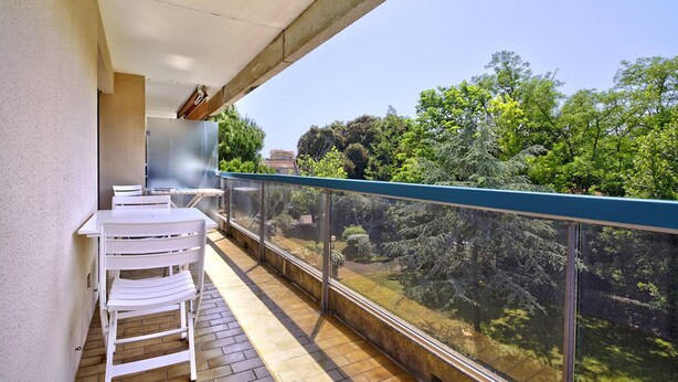 En balkon med et bord og stole, gelænder af glas samt udsigt til træer og bygninger.