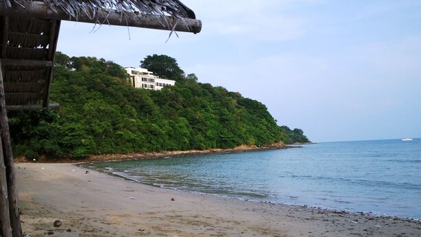 A beach with a thatched-roof structure, a sandy shore, and a building on a hillside. - Summer Breeze Inn Hotel (Wichit)